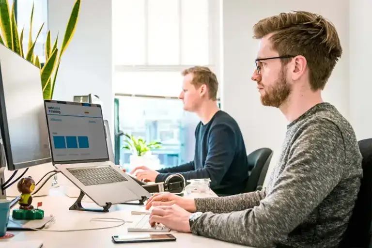Zwei Personen sitzen an einem Schreibtisch. Die Person im Vordergrund hat auf ihrem Bildschirm die Webbased.Training Lernplattform aufgerufen.